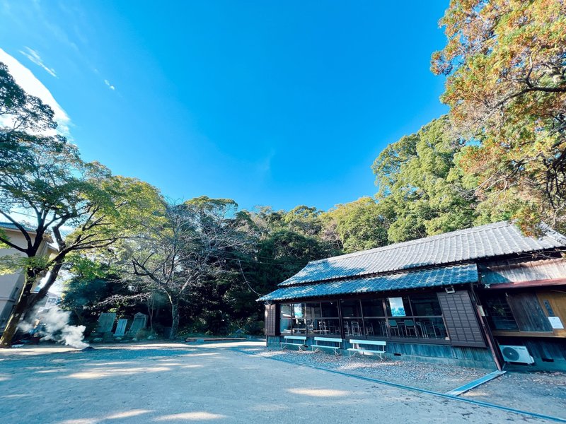 晴れた日の日和佐八幡神社の写真。写真上部半分ほどに青空が写っており、緑色の木々と共に右手前にベンチと建物が見える。