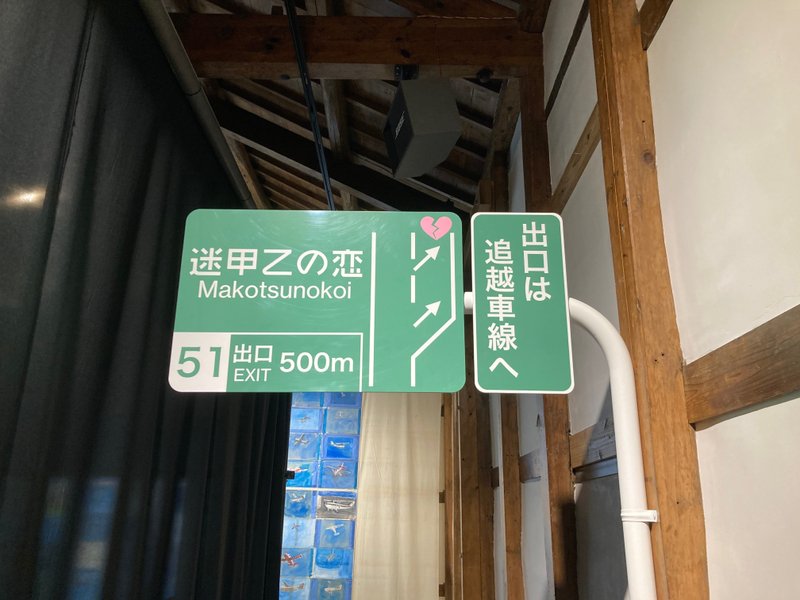 高速道路の降り口のような作品。緑背景に白い文字。「出口は追越車線へ」「迷甲乙の恋Makotsunokoi」「出口EXIT 500m」という文字と出口を示す道路図と矢印があり、出口の先には割れたハートがある。