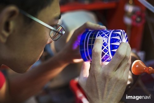A craftsman glass maker holding a vivid blue cut glass cup, inspecting it closely.｜Mint Images