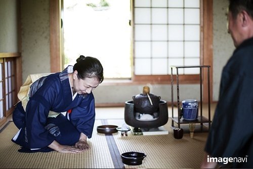  Japanese woman wearing traditional bright blue kimono with cream coloured obi and man kneeling on floor during tea ceremony.｜Mint Images