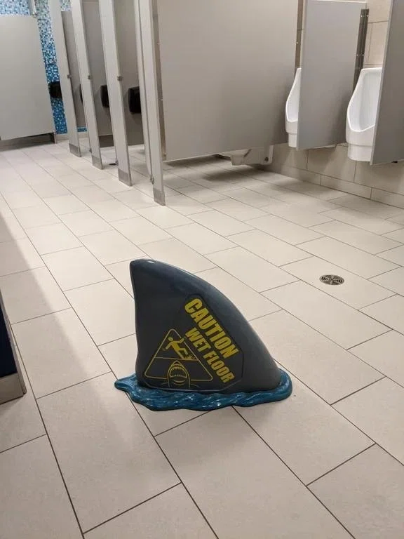 サメのヒレの形をした看板がトイレの床に置いてある。看板には「CAUTION WET FLOOR」の文字