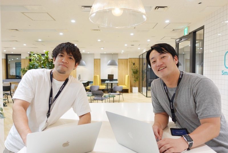 2人の男性（松下さんと山本さん）がそれぞれパソコンを前に置いた状態でカメラに向かって微笑む写真。