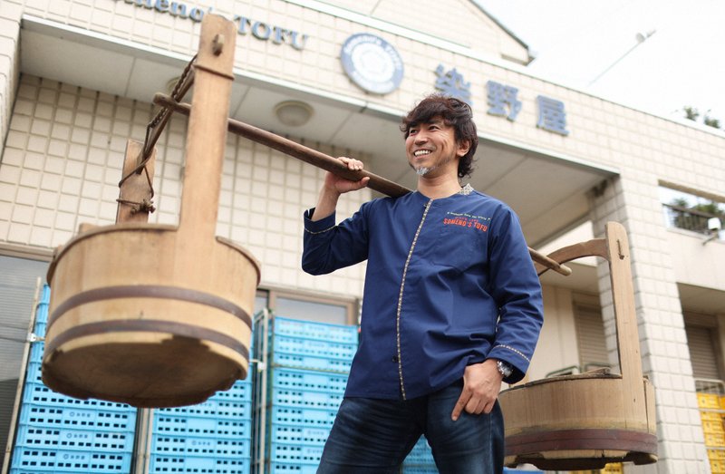 かつて豆腐の販売で使われていたおけを肩に乗せ、ポーズをとる染野屋の小野篤人社長=小川昌宏撮影