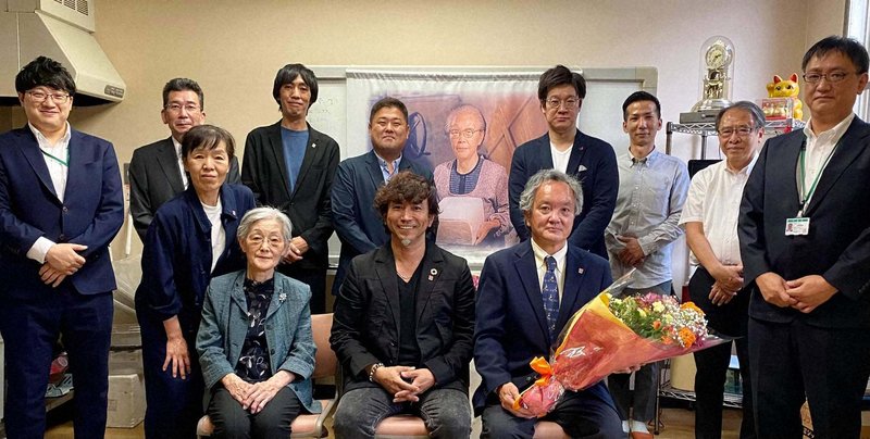 山下ミツ商店の株式譲渡成約式を終え、花束を受け取った山下浩希さん(前列右から2人目)と染野屋の小野篤人さん(同3人目)。背後には祖母ミツさんの肖像が掲げられた=本人提供