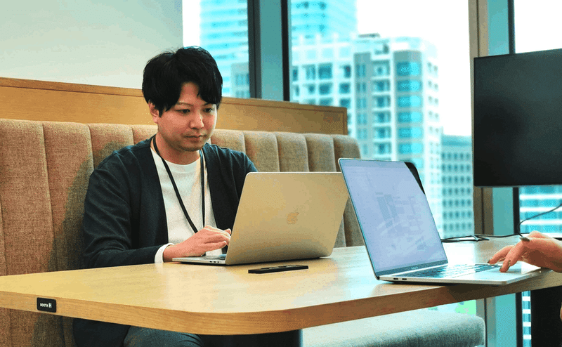 画面左側に写った伊藤浩介さんがパソコンに向かっている写真。画面右手には、向かい合って座っている方のパソコンと手が写っている。