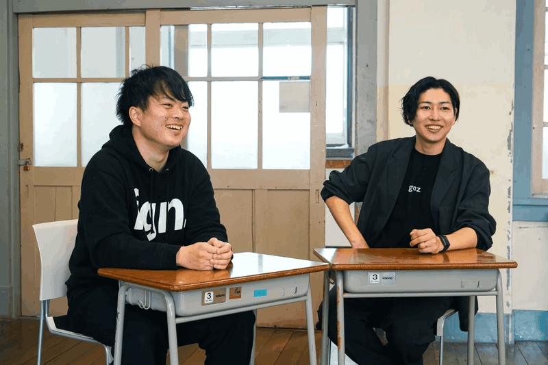小学校で使用される生徒用デスクを前に、椅子に腰掛ける、笑顔の崎山さんと、笑顔の吉岡さん