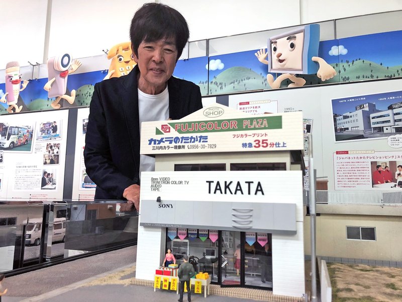 「ジャパネットたかた」のスタート地点になったカメラ店のミニチュア模型をのぞきこむ同社創業者の髙田明さん=清水憲司撮影