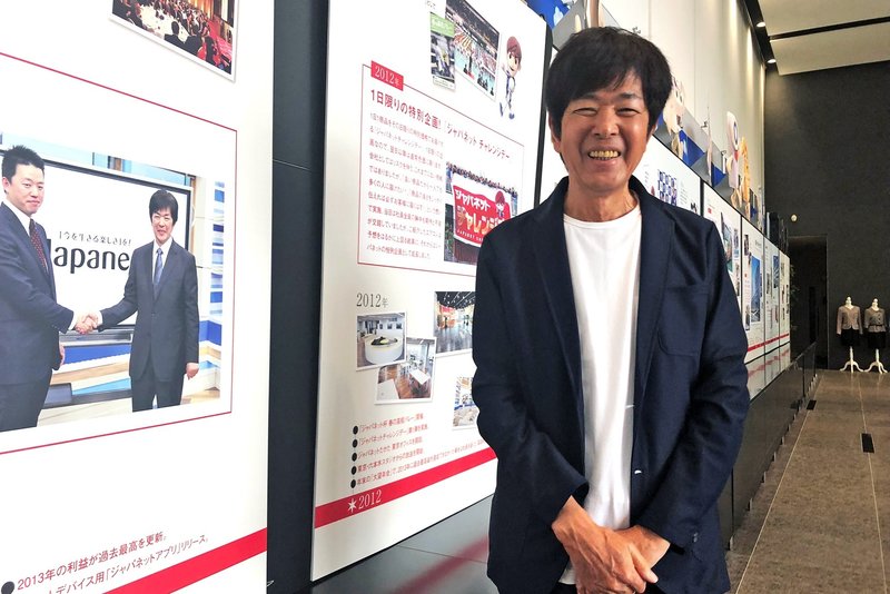 ジャパネットたかた社長の髙田旭人さんと握手する写真の前で笑顔を見せる同社創業者の髙田明さん=清水憲司撮影