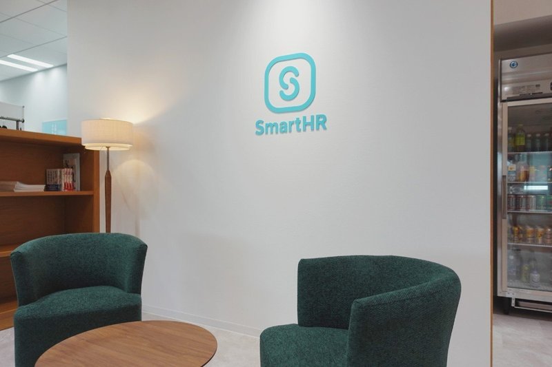 キャプションの通りのオフィスの様子。写真中央には白い壁と「SmartHR」というロゴとアイコン、その下には緑色のソファーが2脚ある。左奥には本棚と間接照明が見える。