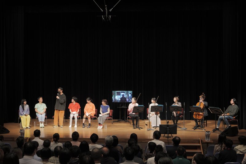 舞台左側に椅子に座る俳優５名とマイクを持つ岡田、中央にモニターに映し出される藤倉、右側に椅子に座る日本の演奏者５名