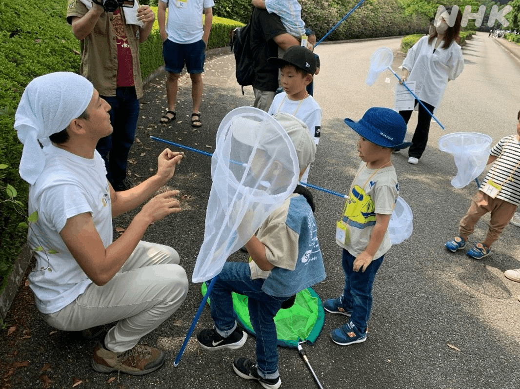 屋外で網を持った子どもたちに囲まれながら、左手に持ったセミを説明する宮﨑アナ。