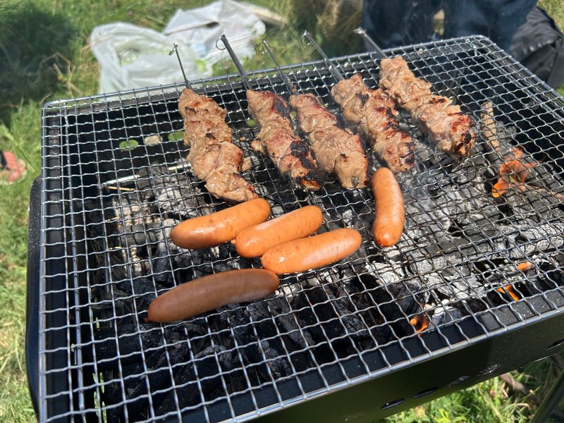 BBQコンロの網の上で鉄串に刺された羊肉とソーセージが焼かれている写真。