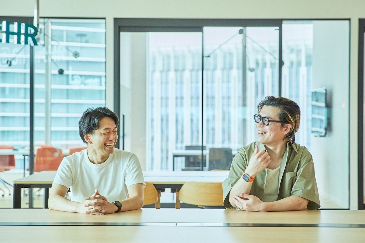 机を前に並んで座り、向き合って大きく笑っている男性2人の写真。