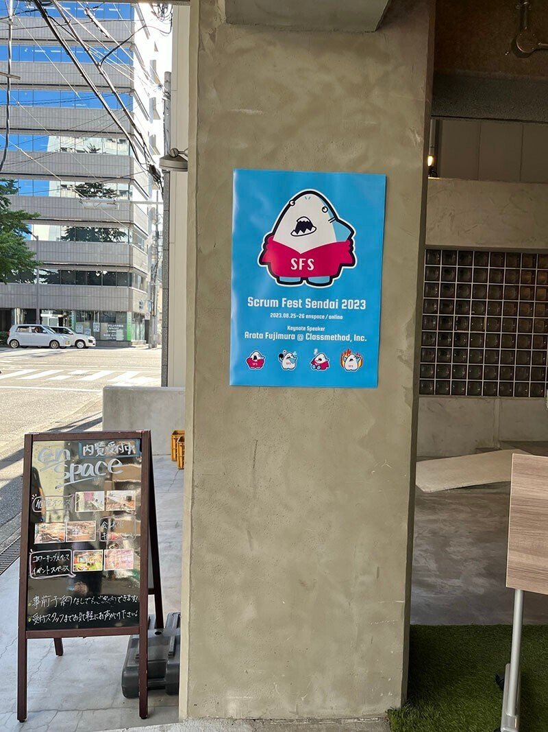 enspaceの入り口の柱に水色の背景に濃いピンクのパーカーを着たシャークレ君があしらわれたScrum Fest Sendai 2023のポスターが貼られている