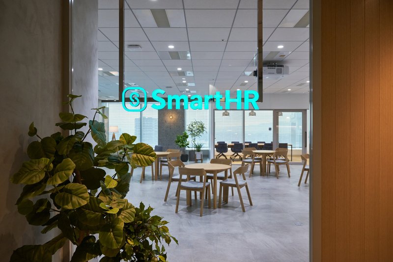「SmartHR」という光る文字がぶら下がっており、手前左には木が、奥には椅子と机が並んでいる様子が見える写真。