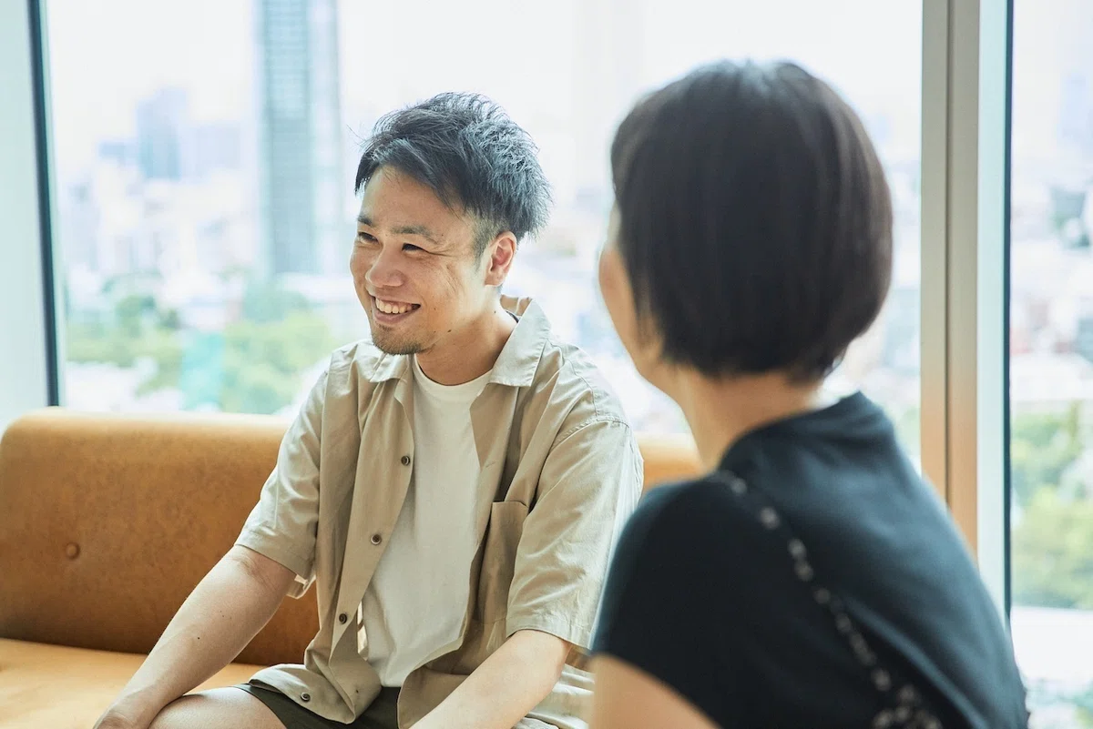 左側を向き、微笑む男性の写真。右手前には女性の後頭部が写っている。