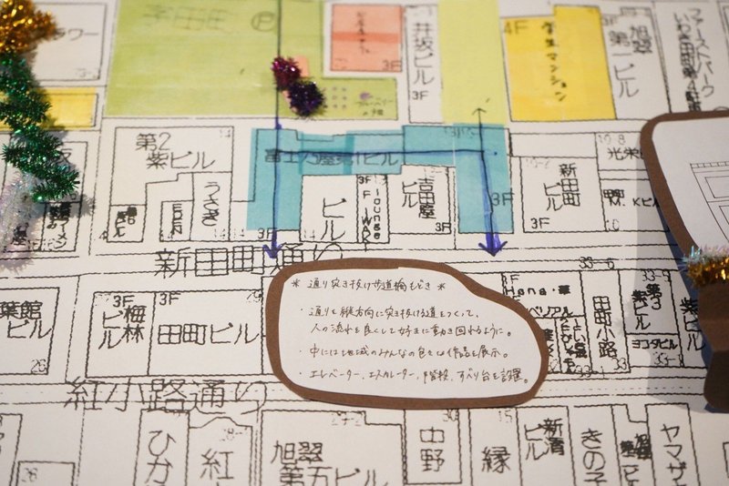 マップの拡大写真　水色のマスキングテープがコの字型に貼られておりその上に青色のペンで矢印が書かれている　その下に、画用紙で作られた吹き出しに通り抜けトンネルについての説明が書かれている
