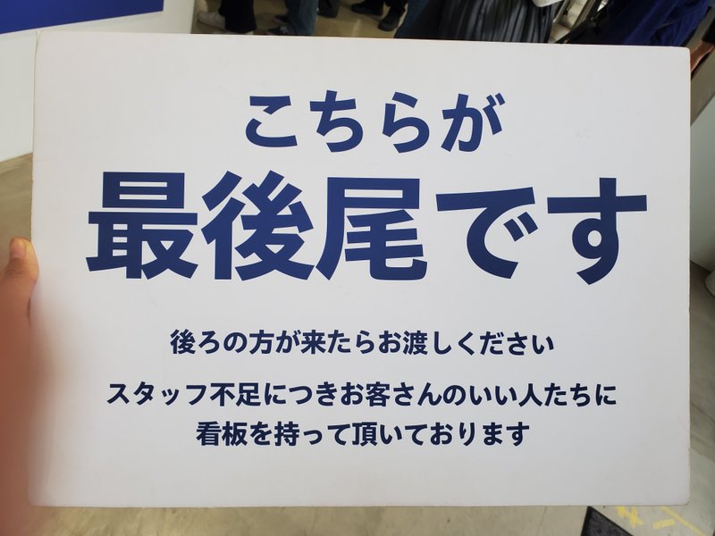 「こちらが最後尾です　後ろの方が来たらお渡しください　スタッフ不足につきお客さんのいい人たちに看板を持って頂いております」と書いてある看板。