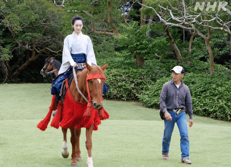 衣裳を着た愛希さんが、赤い衣裳を着た馬にまたがりこちらに向かってくるとなりで、見守りながら歩いてくる馬術スタッフ