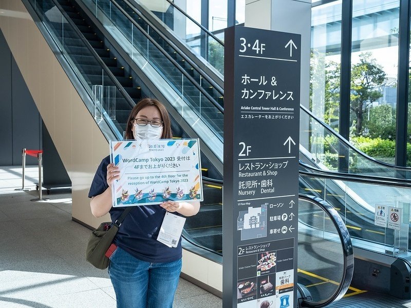 エスカレーターの隣で「WordCamp Tokyo 2023 受付は4階までお上がりください」のボードを掲げている笑顔のyshimizu