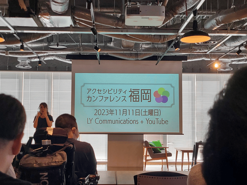 写真 アクセシビリティカンファレンスのタイトル、ロゴが映っているスライドを写している。隣には手話通訳の女性