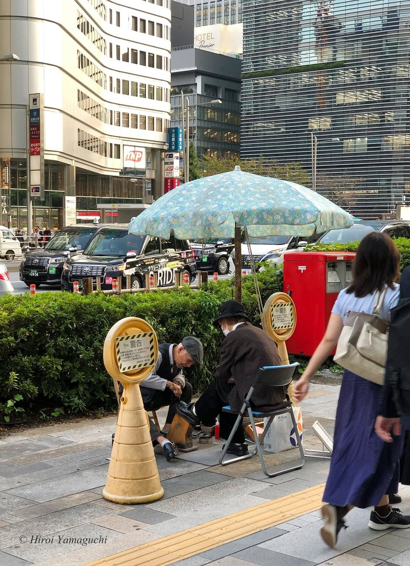 靴磨きの職人さんが年配の女性の黒い靴を磨いています。日除の傘もお手製の様子。