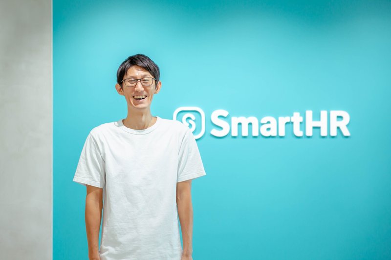 SmartHRのロゴを背景に、笑顔で写る廣辻さん