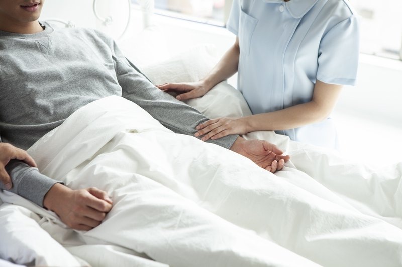 病院のベッドに横たわる男性と、そばで腕にそっと触れる看護師のイメージ画像