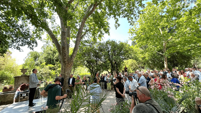 木々のなか、式典で話す人を見守る大勢の人達の写真