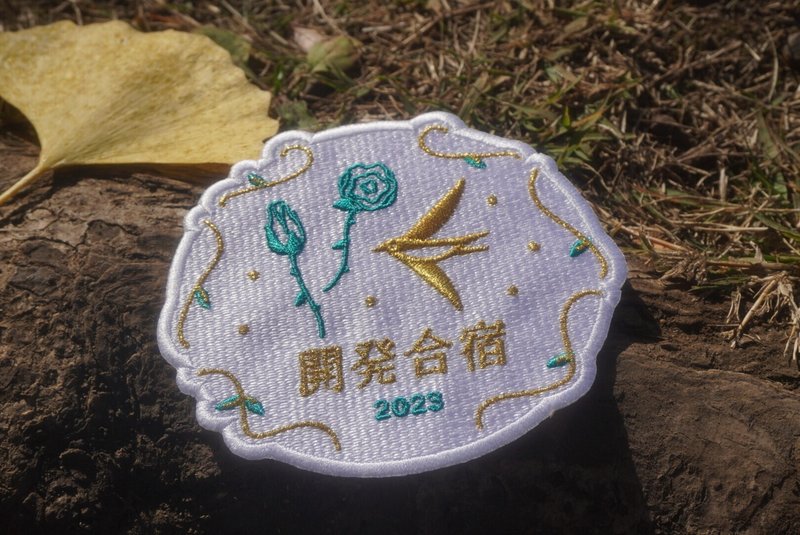 キービジュアルのロゴワッペン。金色と緑の糸で刺繍されている。freeeのツバメやバラ、「開発合宿2023」の文字が刺繍されている