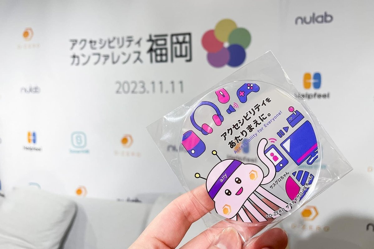 アクセシビリティカンファレンス福岡 ロゴをバックに、「アクセシビリティをあたりまえに。」のテキスト入りアクリルコースターが映っている写真