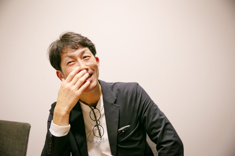 北村さんのバストアップの写真。笑顔を浮かべ、口元に片手を寄せている。