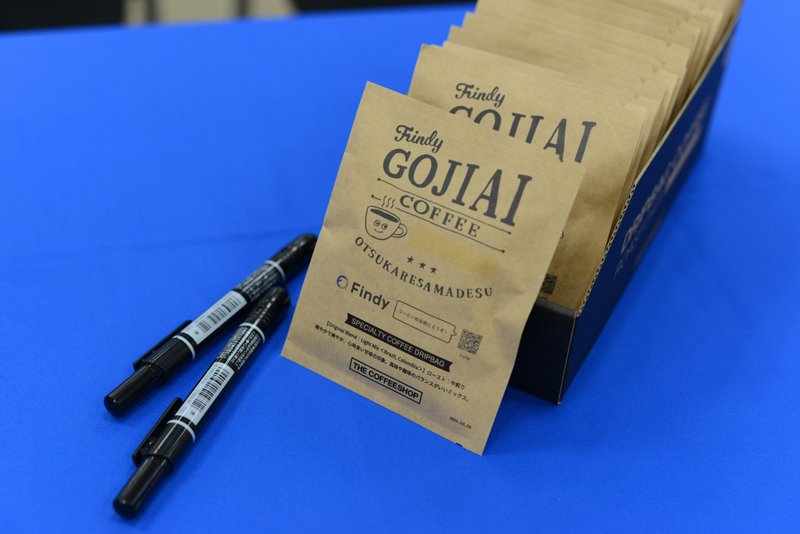 「Findy GOJIAI COFFEE」と書かれた薄茶色の紙で包装されたコーヒーが並んでいる。下の台には青色の布が敷かれており、左側には2本のサインペンが置かれている。