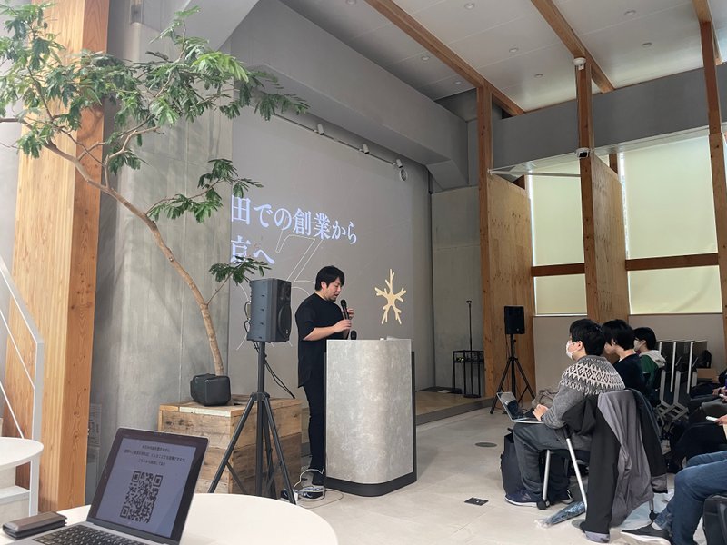 イベント会場で阿部 文人（あべ・ふみと）さんがマイクを持ちながら話している。スライドには「田舎での創業から東京へ」と書かれている。