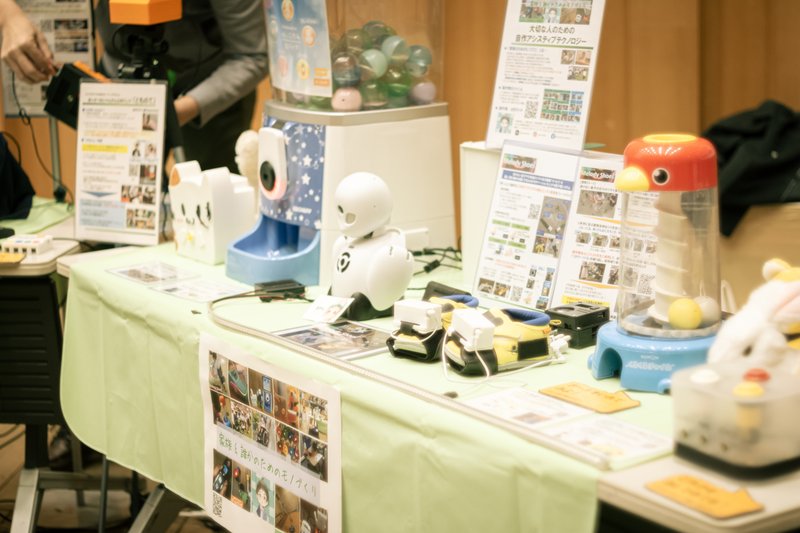 「OGIMOテック研究室」の展示品を並べた写真。画面上には手をかざすことでアイテムが出てくるガシャポンや、分身ロボット「OriHime」といったロボットが展示されている。