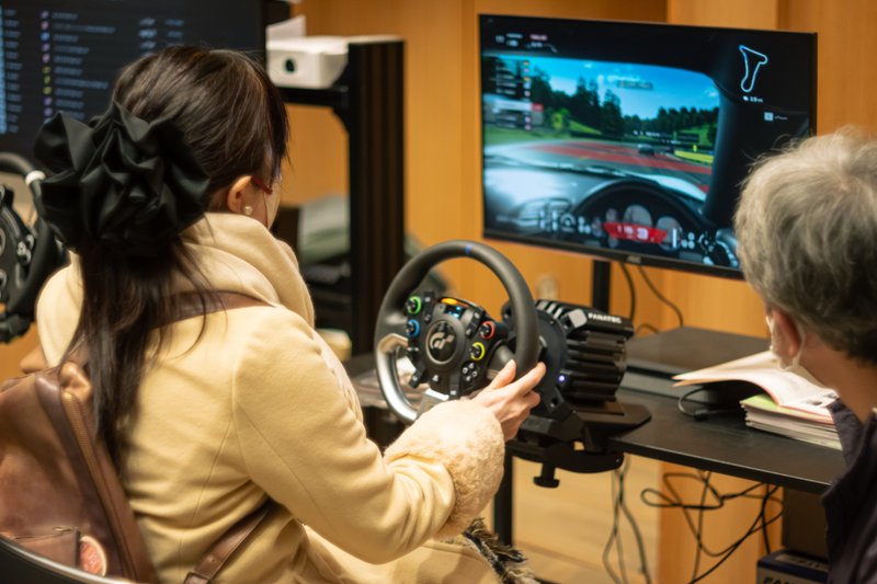 eレースゲームで使用するハンドルコントローラーを手のみで操作できるよう作られた装置を、体験者が操作している写真
