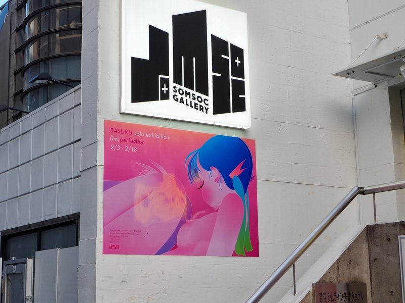白いモルタル壁の建物に、「SOMSOC GALLERY」の大きな看板と、その下にはピンクのカラーが印象的な少女のイラストを大きく載せた「RASUKU solo exhibition （im)perfections」のポスターが貼られている