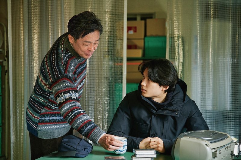 場面写真　作業場でセーターを着た栗田社長が黒い上着を着た山添くんにお茶を出す