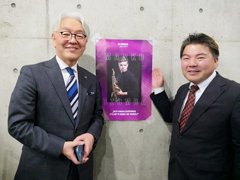 埼玉県富士見市 鶴瀬コミュニティーセンターで、セルビア共和国出身の国際的に活躍するソリストのマルコ・ジョンバ（サックス）さんのポスターを挟んで、スーツを着た星野光弘市長とスーツを着た榊原平さんの3人が立っている光景