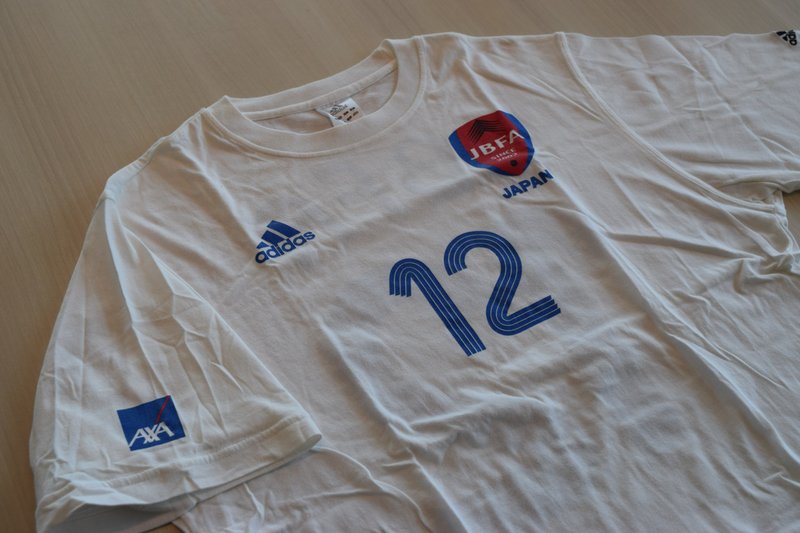 白いTシャツの真ん中に、サポーターの番号である12番の数字が印字されている。また右肩にアクサのロゴ、右胸にアディダスロゴ、左胸にJBFAロゴが印字されている。