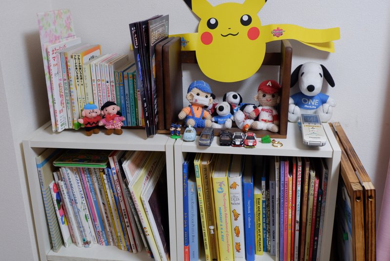 白い本棚の写真。英語や日本語で書かれた絵本が並ぶ。棚の上には手作りの人形や、ミニカー、紙に印刷されたピカチュウなどが本と一緒に置かれている。