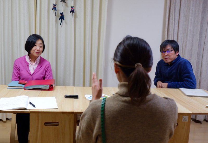 インタビューの様子。中央にはれもんハウスを運営する藤田さんが手でジェスチャーしながら話している後ろ姿。それを机の向こうから聞いている佐々木夫婦。