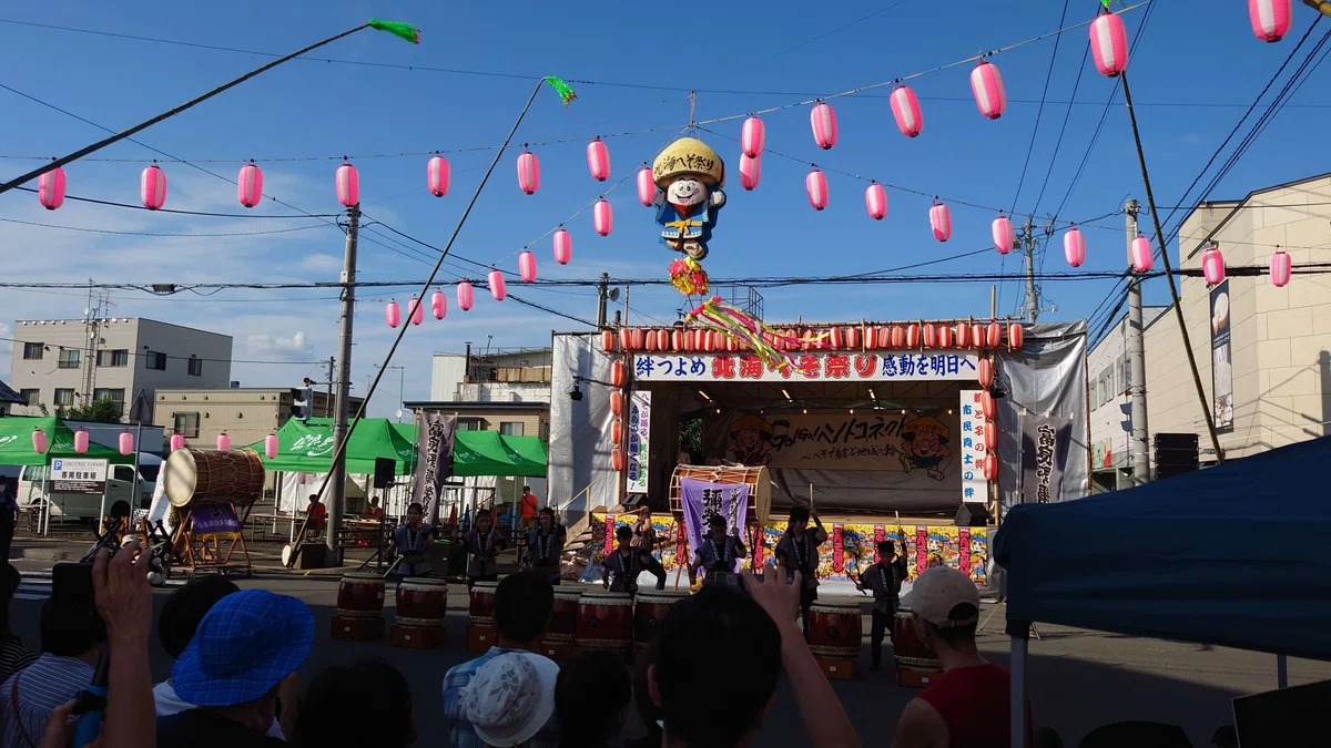 お祭り会場にたくさんの提灯がぶら下げられ,ステージの前で和太鼓を演奏する人たちが並んでいる.