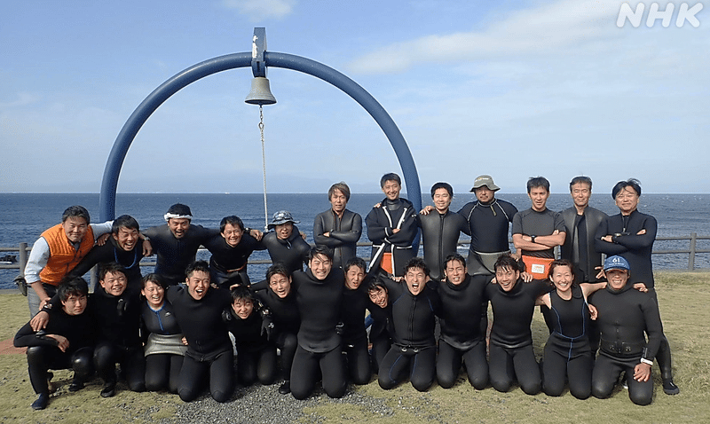 海をバックに、ウェットスーツを着た潜水班26人が並んだ集合写真。