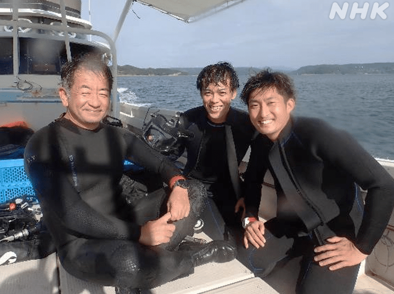 海上の船のうえでウェットスーツ姿でカメラににっこりする３人。