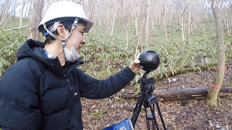 熊笹の生い茂る山中で、ヘルメットをかぶり丸いVRカメラを触る田中さんの写真。