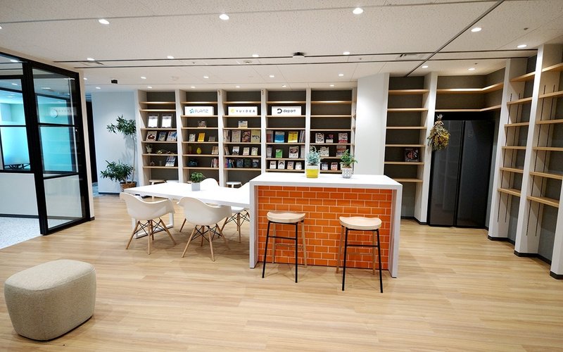 写真：エントランス/中央にちっきんカウンターのようなテーブルと椅子が並び壁一面に本棚がある