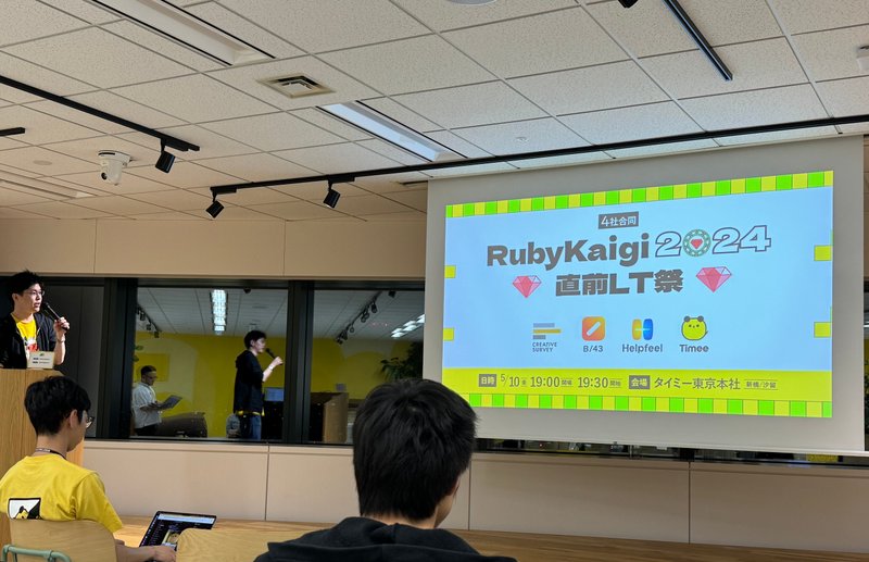 室内でイベントが開催されている様子の写真です。スクリーンには「RubyKaigi 2024 直前LT祭」と表示されています。左手側に立っている司会の男性は黄色いTシャツを着ています。