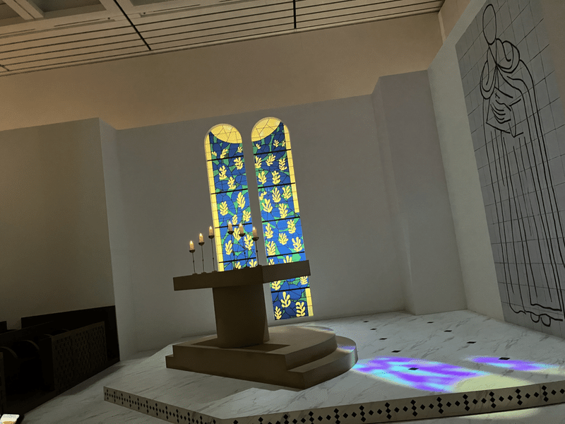 礼拝堂のスペース。黄色と青のステンドグラスに、蝋燭がたった祭壇みたいなところ。壁は白くて、右側には大きな神職者の絵が描いてある