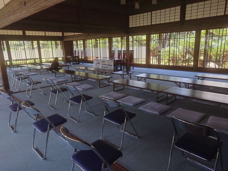 窓の向こうに日本庭園が覗く，パイプ椅子と座布団と長机が並べられた広間の中心に「PHP Conference Kagawa」の幕が張られた演題と椅子があり，広間の中央左に一人の人が座っています．